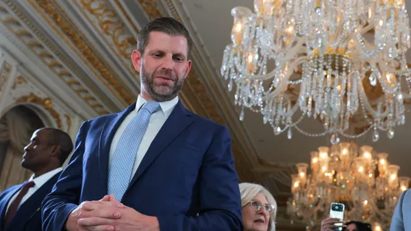 A man with a five o'clock shadow and wearing a three-piece blue suit looking down, with a chandelier in the background.