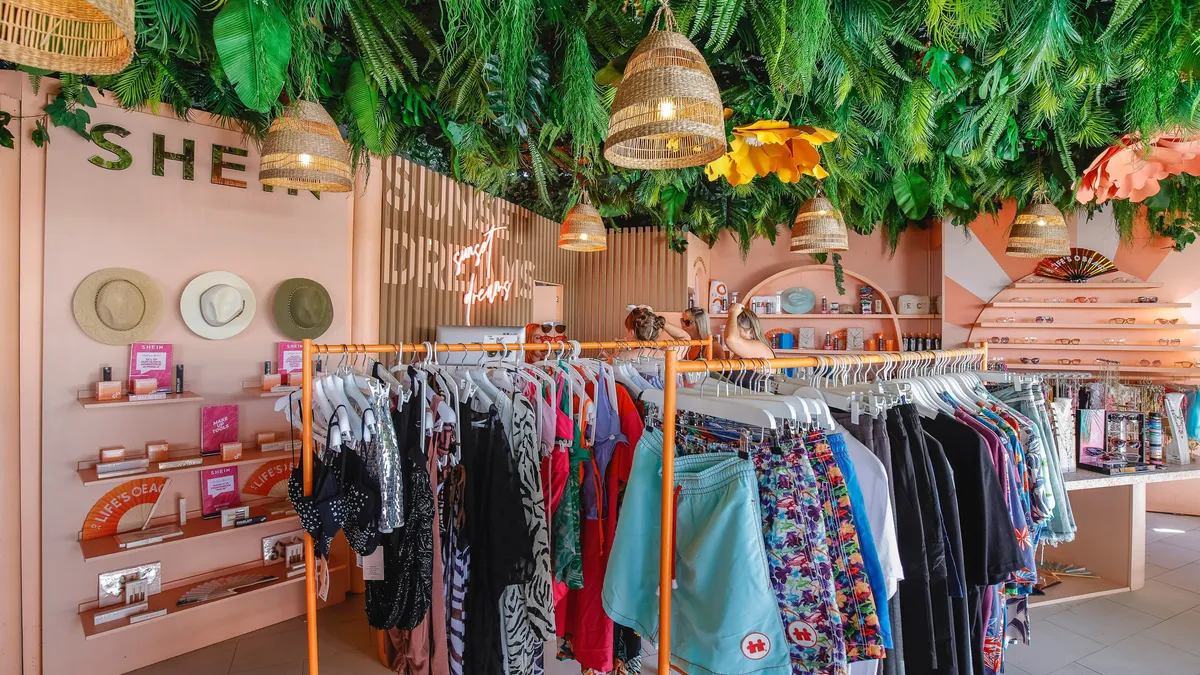 A general view of a Shein fashion show & pop-up shop at O Beach Ibiza. Palm fronds on the ceiling, pink walls with hats displayed, and racks of clothing in the store.