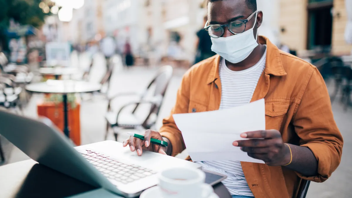 African American male student sitting in an outdoor cafe wearing a protective face mask, studying using a book and a laptop