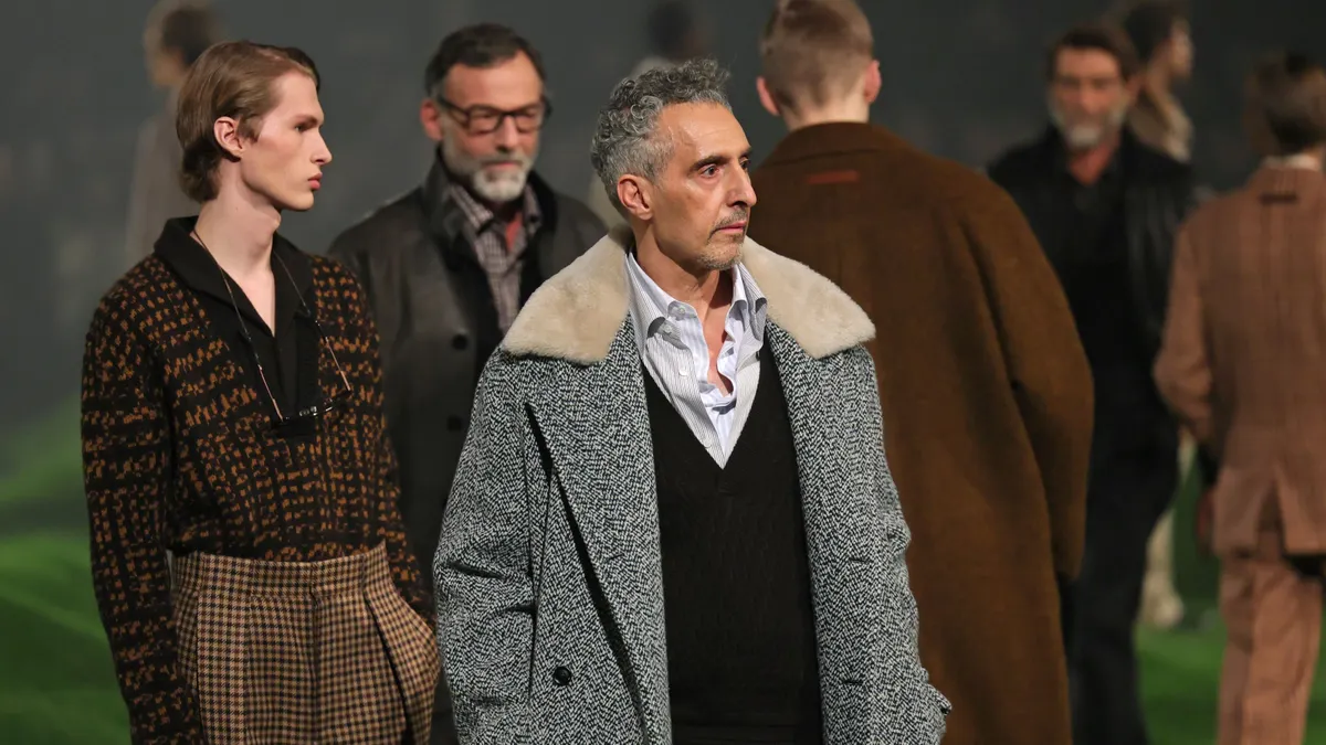 A man in a gray coat stands in front of multiple models on a fashion runway.