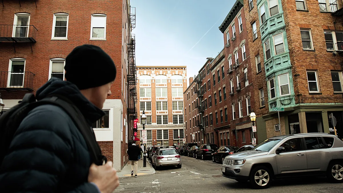 A person ina black puffy jacket, black beanie and backpack walks along a street with residential brick buildings and parked cars.