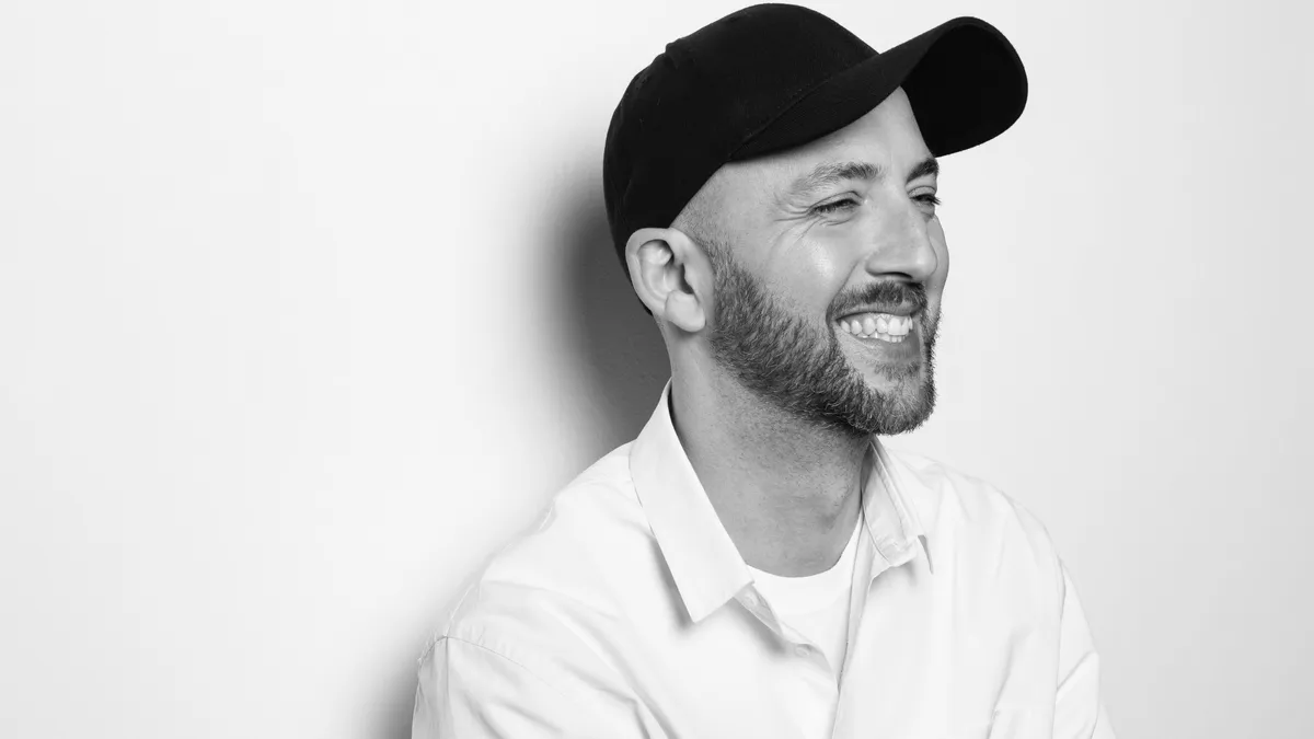 A person wearing a white shirt, white t-shirt and black baseball cap leans against a white background smiling.