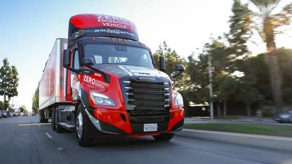 A battery electric Reyes Coca-Cola Bottling truck travels down a road with trees in the background.