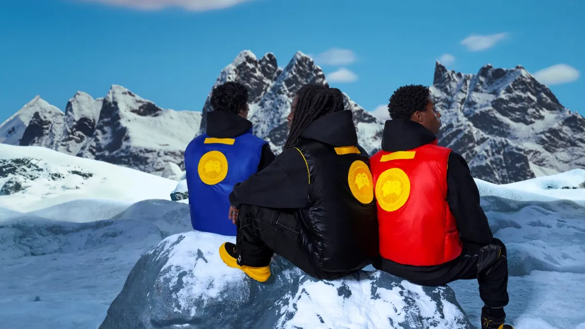 Three men sit on a snowy landscape wearing boots and quilted Canada Goose x Pyer Moss collection vests in blue, black and red.