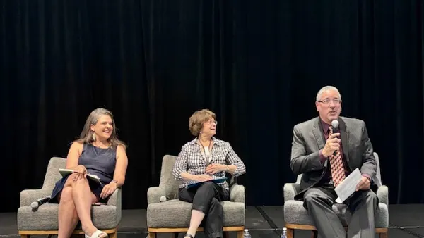 Three people are sitting in chairs on a stage. One person has a microphone