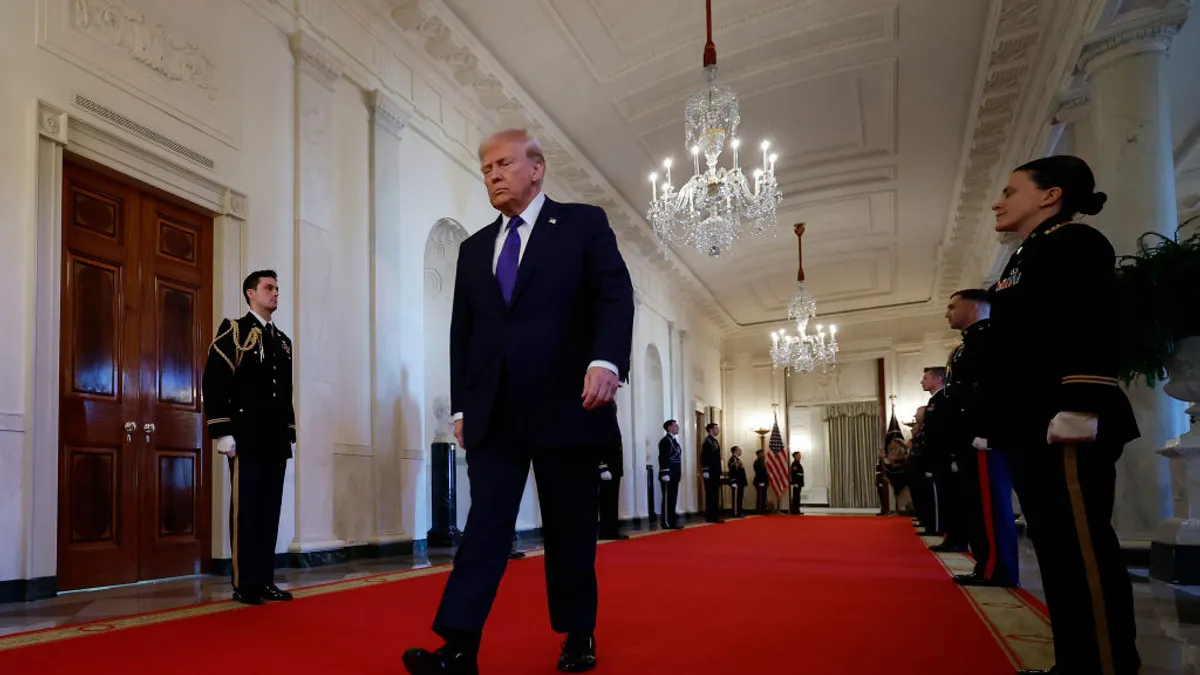 Donald Trump is walking in a hallway of the White House. The photo is taken from the ground up. There is red carpet and people in uniform are lined along the walls of the hallway