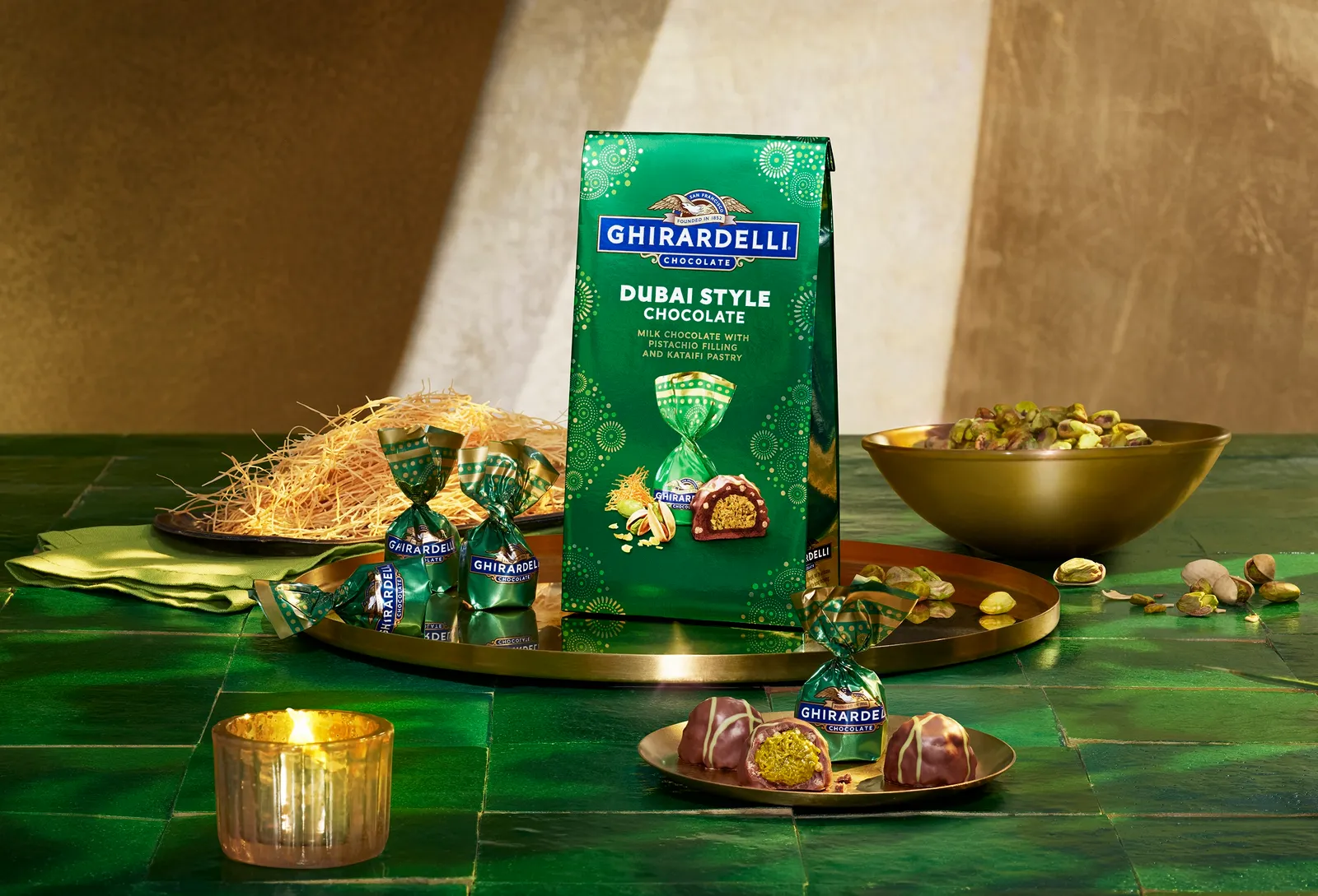 A bag of candy sits on a green table along with a bowl of pistachios, with several wrapped candies and nuts scattered around. The bag of candy says Ghirardelli chocolate Dubai-style chocolate. Milk chocolate with pistachio filling and kataifi pastry.