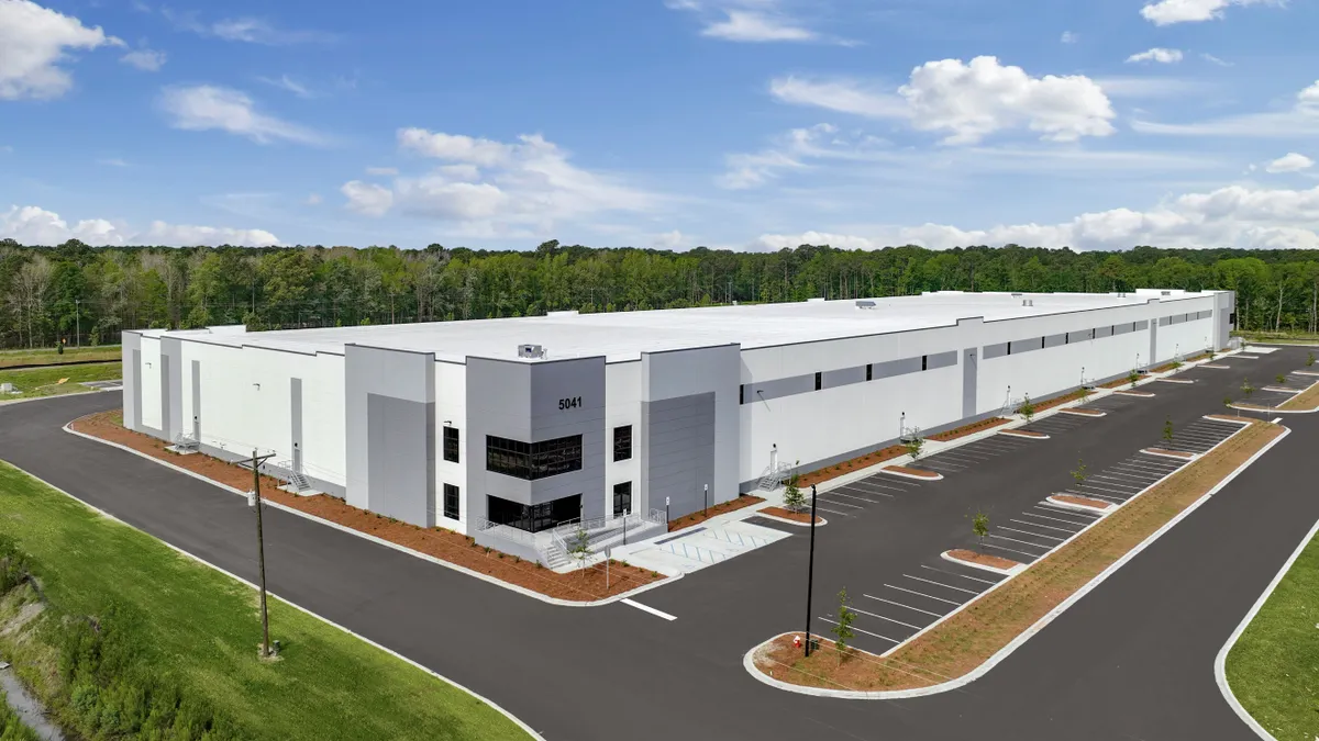 A white building surrounded by a dark gray parking lot and green landscape under a blue sky with white clouds during the day.