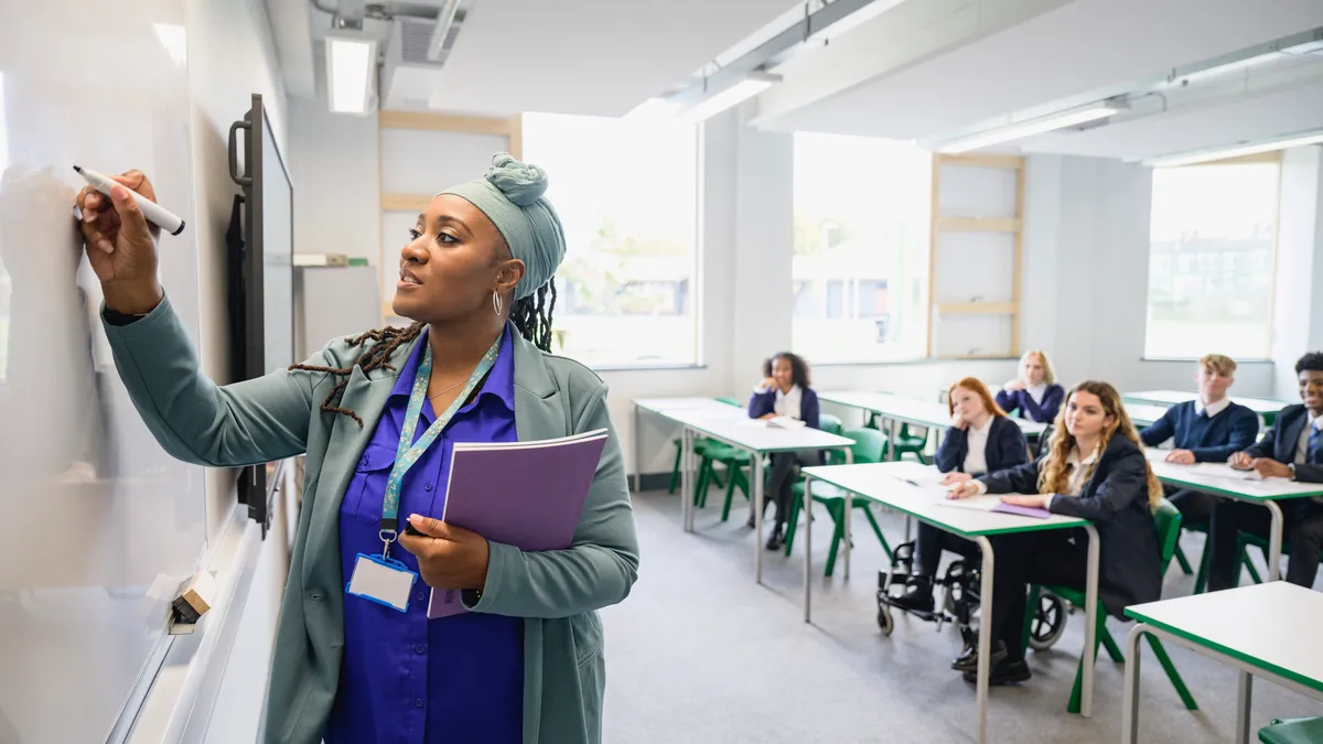 Side view of female educator teaching class as teenage students in uniforms sit at desks in background watching and listening.