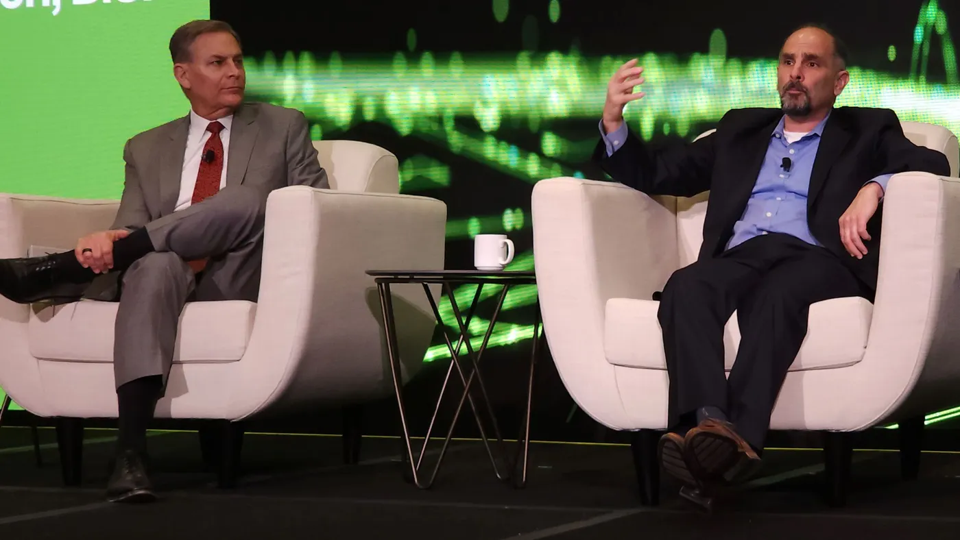 Two men in business suits sitting in white chairs with a black and bright green screen in the background.