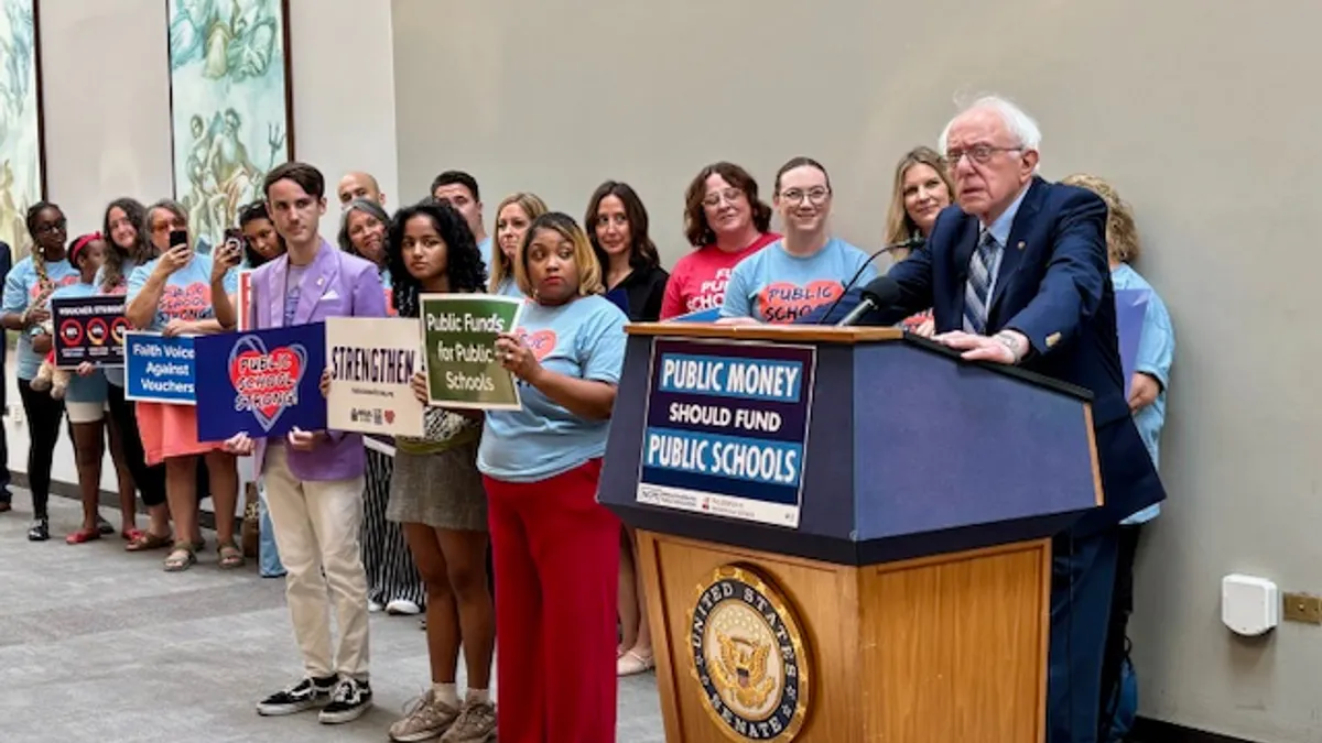 A person is standing at a podium with a microphone inside a large room. Behind the speaker are other people holding signs supporting public schools.