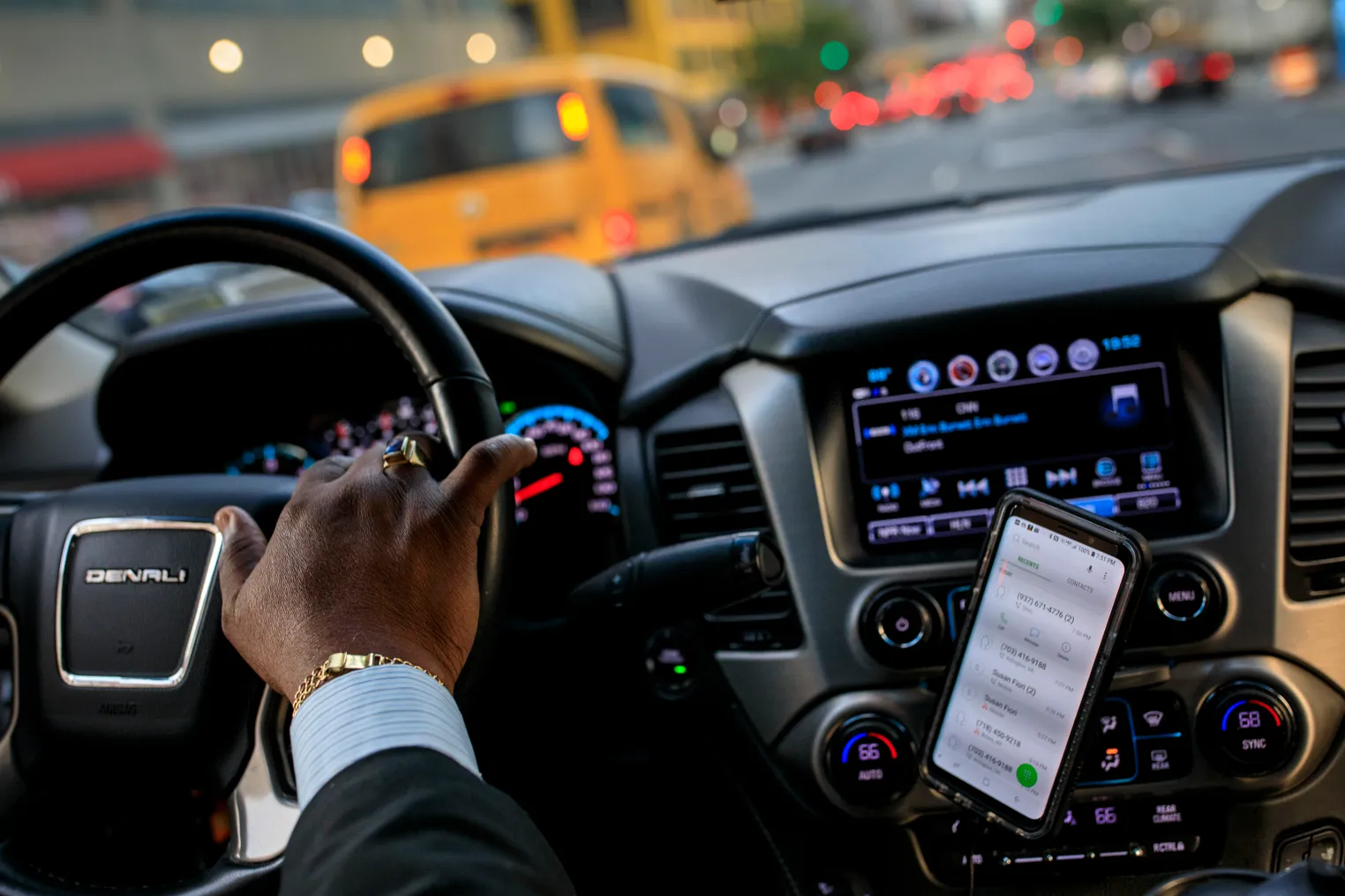 Car driver's hand on the wheel with a smartphone to the right.