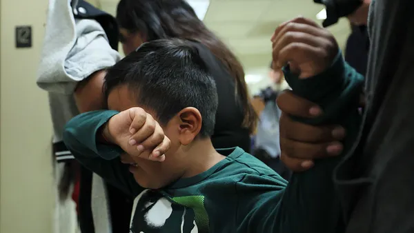 A child is holding their arm over their face. Their other arm is being held by someone.