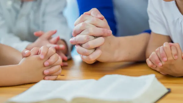A close up of clasped hands clasped in prayer are pictured around a table with an open bible.