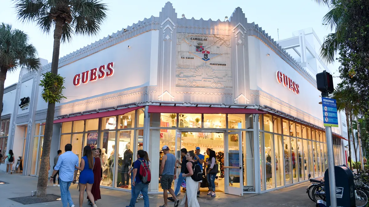 Outside corner view of a Guess store, with a white facade, palm trees lining the street, and crowds of shoppers milling about.