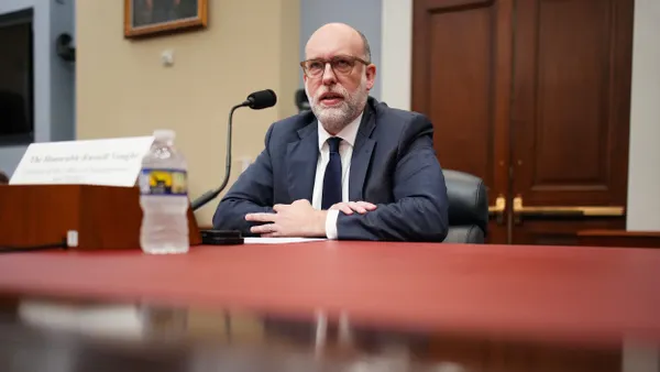 A bald man in a dark suit and tie sits at a desk in front of a microphone.