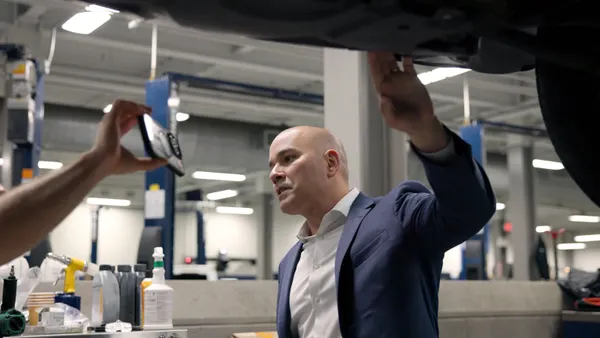 A man talks to someone holding a phone up horizontally, as his left hand reaches up to touch the floor of a vehicle, which is elevated several feet off the ground for service operations.