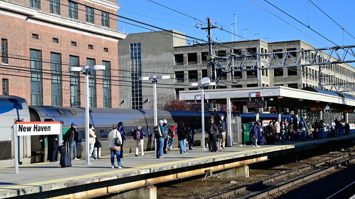Many people walking and standing on a train station platform alongside a train with buildings in the background.