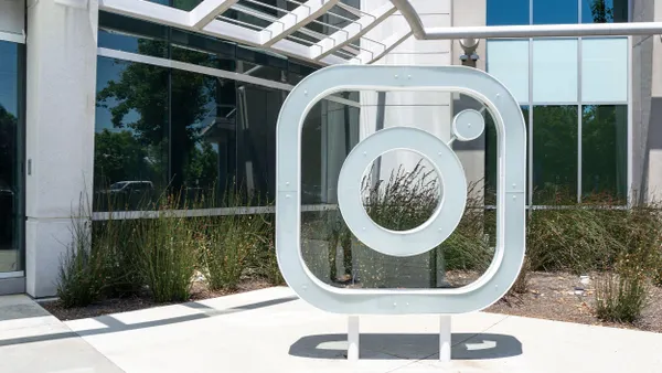 A large, freestanding white sculpture shaped like the Instagram logo is placed outdoors in front of a modern glass building. Surrounding the sculpture are plants and a light-colored concrete walkway.