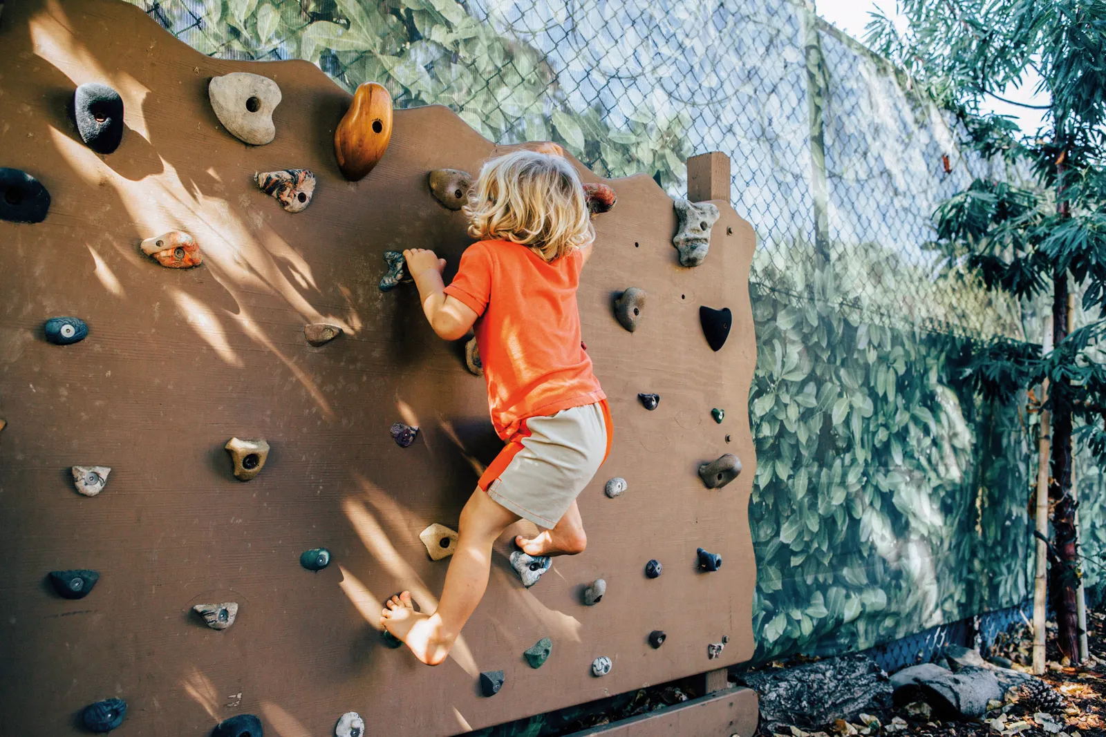 A young child in an orange shirt and beige shorts hangs on a rock wall.