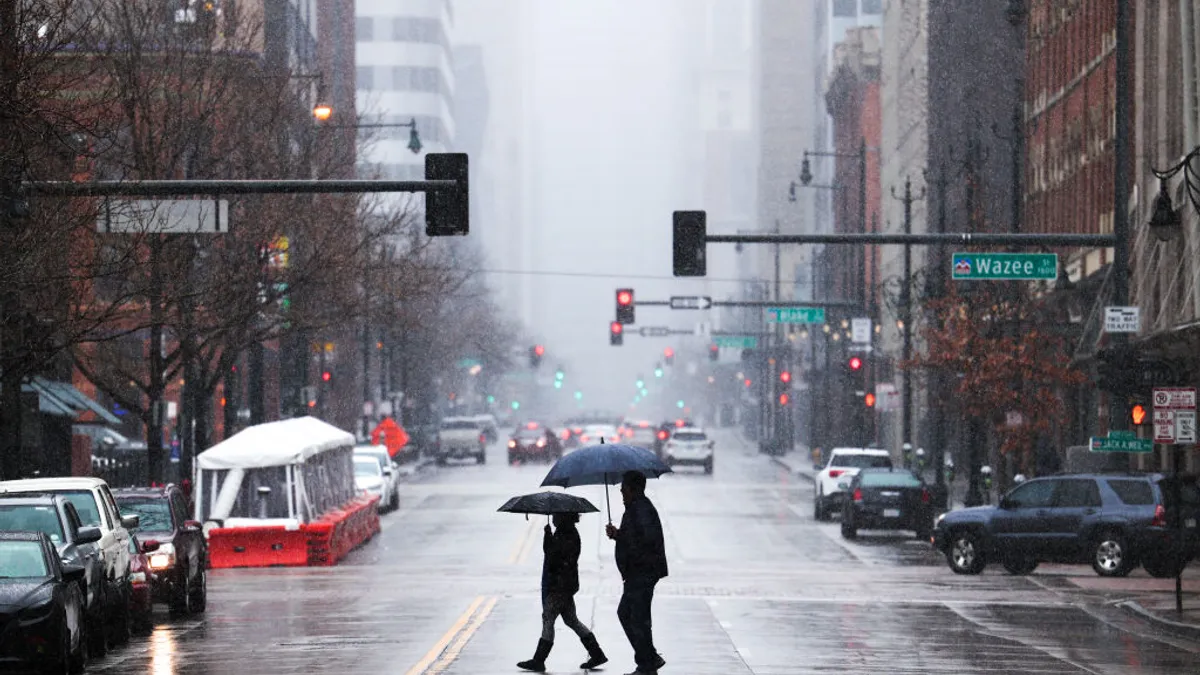 Two people holding umbrellas walk across a city street lined with cars.