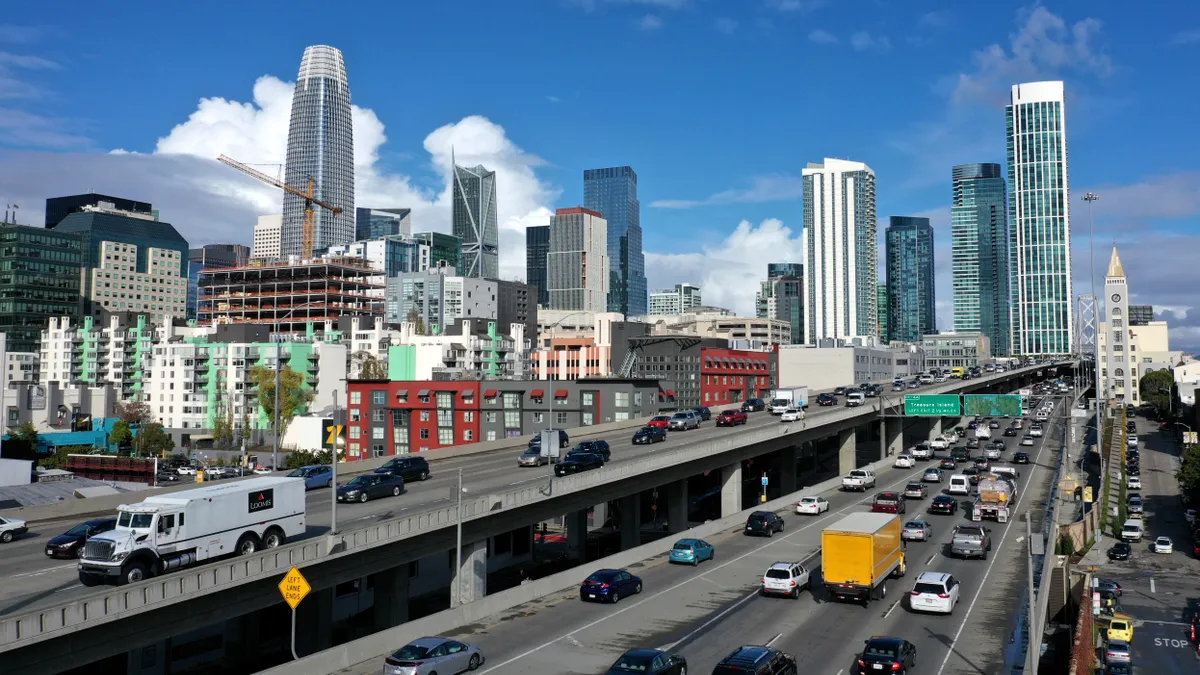 Traffic moves along a highway in San Francisco, California, with buildings and skyscrapers in the background, under a blue sky.