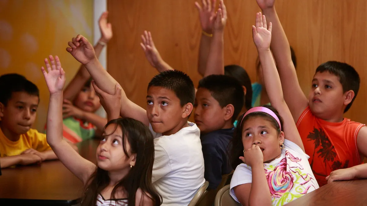 School-age children sit around tables and raise their hands