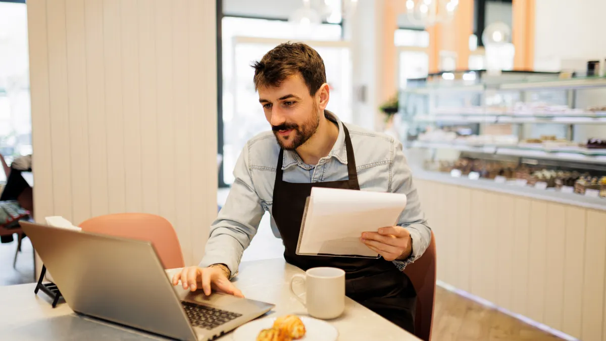 Smiling young male cakes shop owner working a laptop while sitting at a table in his artisanal shop