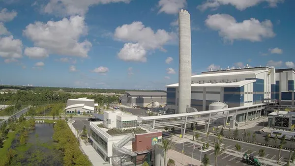 A multi-story production facility with a large, tall smokestack depicted in a picture taken from the air on a sunny day.