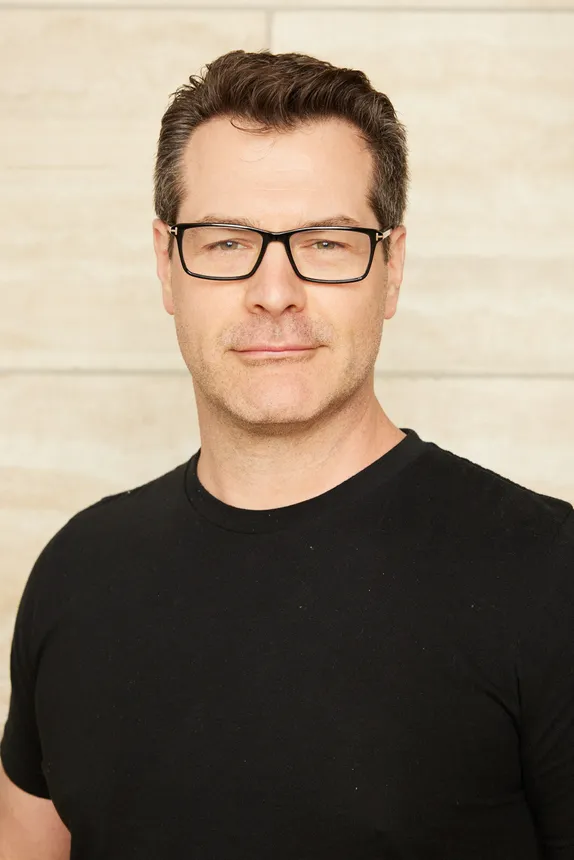 A person with short hair, glasses and a light beard wears a short-sleeved black T-shirt.