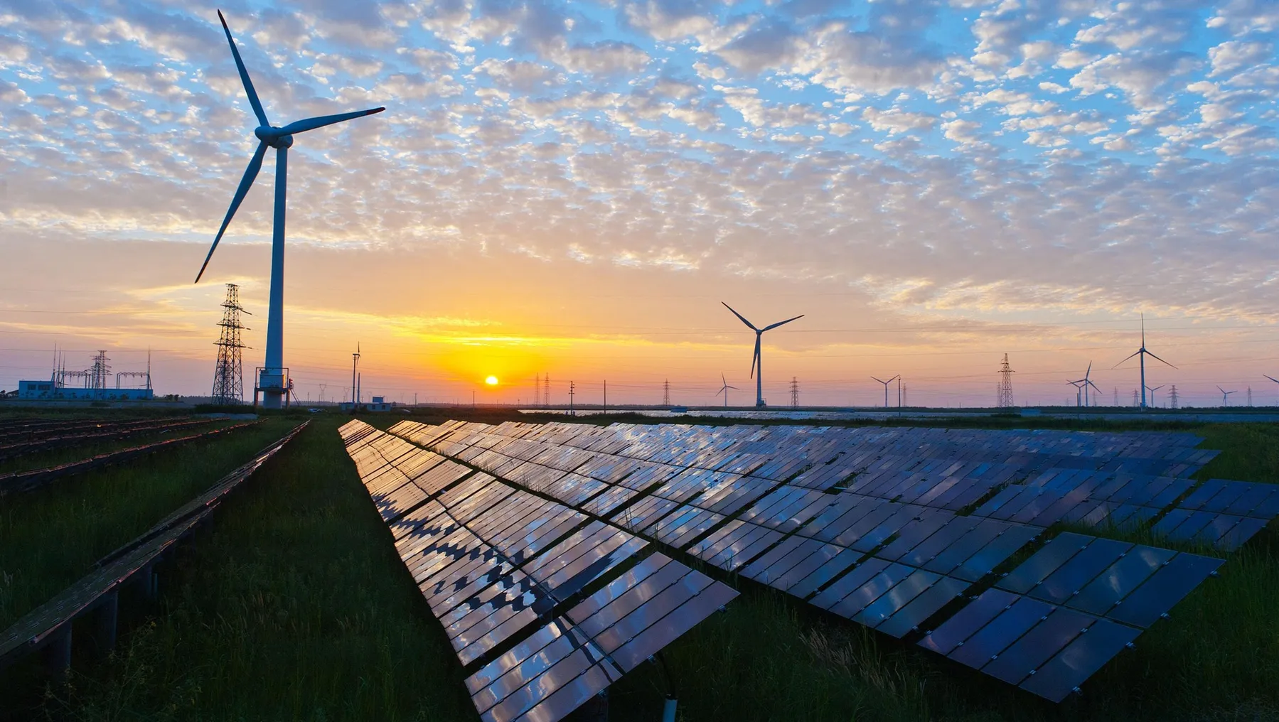 Solar cell panels in the foreground, wind turbines in the middle ground, and electricity pylons in the background