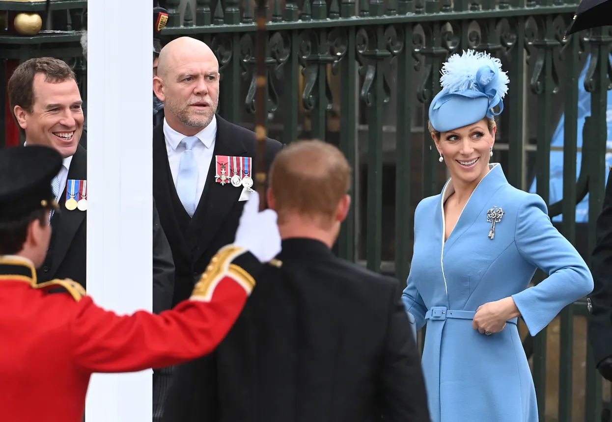 A woman in a blue coatdress and blue fascinator stands among a group of men in suits.