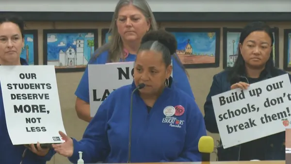 A person stands before a microphone with three others standing behind holding signs that say "Our students deserve more, not less" and "Build up our schools, don't break them!"