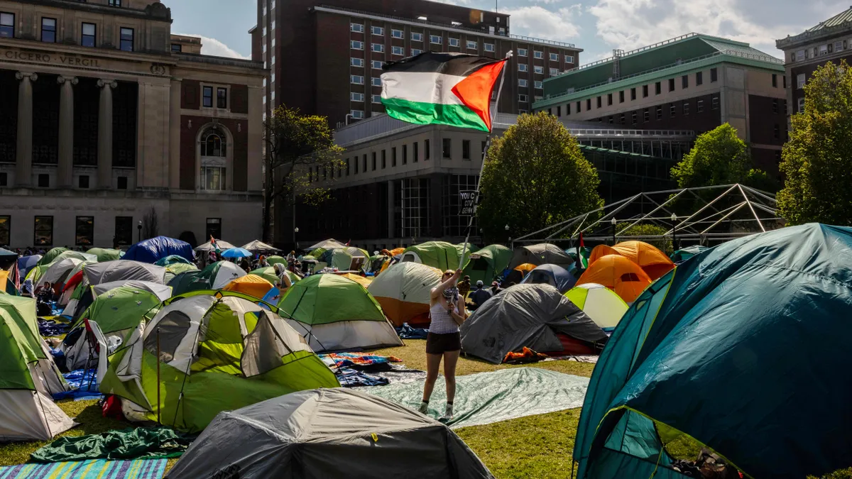 A person standing in the middle of multiple tents holds a flag, the background is a skyline of buildings.