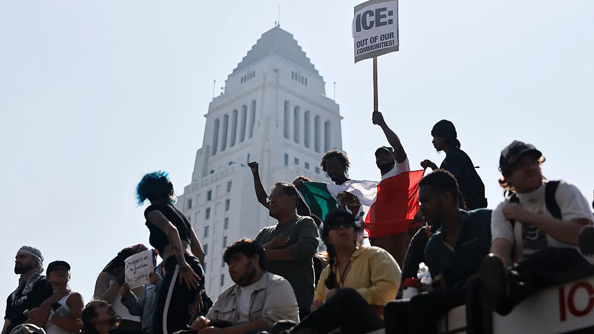 A group of people sit on a bridge in front of a tall white building, one holding a sign that says "ICE out of our communities."