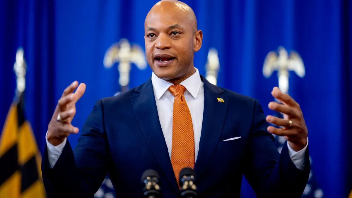 Gov. Wes Moore speaks while raising his hands at a podium in front of a blue background.