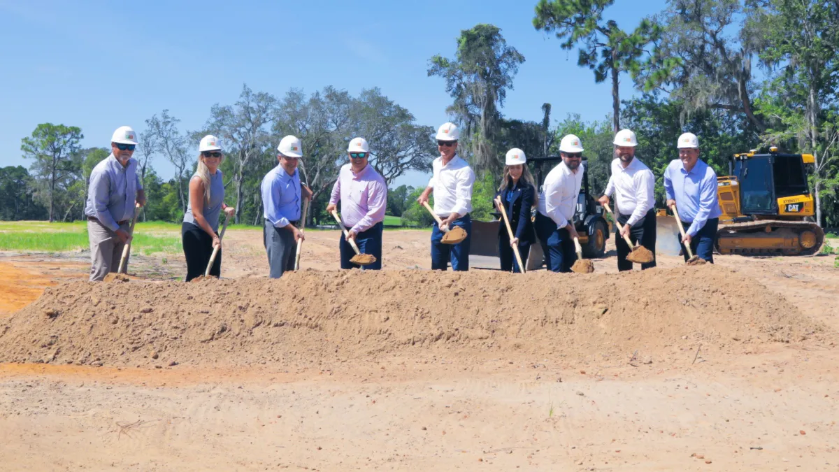 Nine people in white hard hats hold shovels full of dirt in front of a dirt pile.