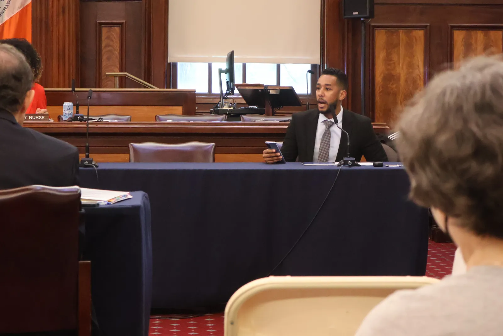 A man wearing a suit sits at a table while holding up a phone and speaking toward people facing him.