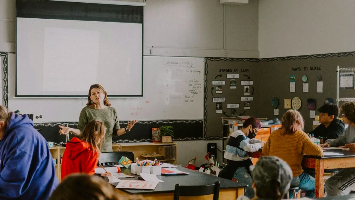 Students are sitting at desks in a classroom. An adult is standing.