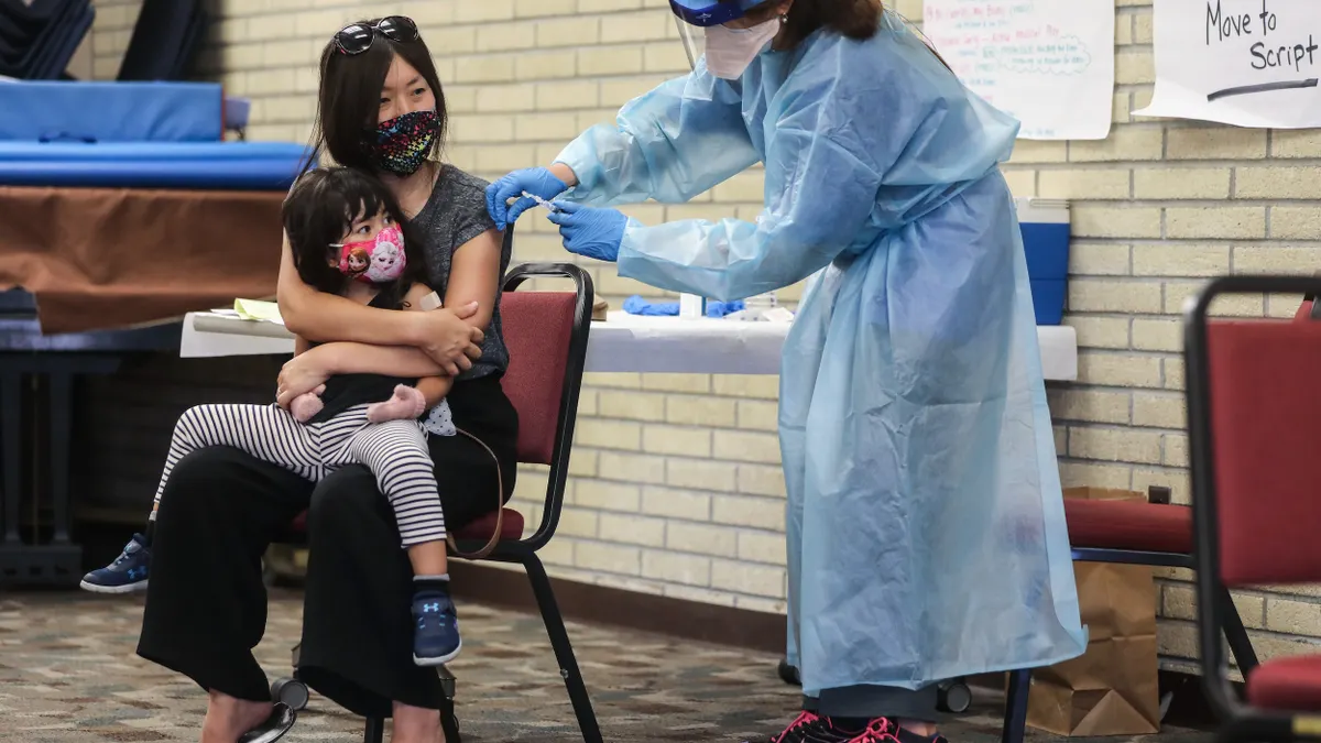 Person wearing mask sitting in chair with toddler in lap receives a shot from a person wearing mask and medical gown.