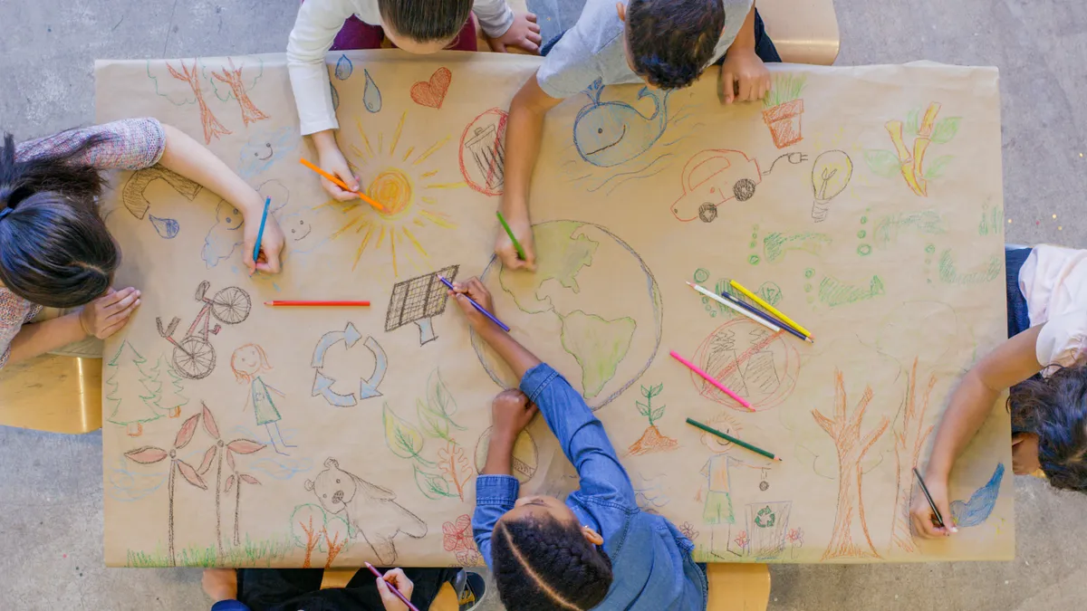 An aerial view of students sitting around a table that is covered with paper. The students are drawing images on the paper of a globe, car, bike, plants, and more.