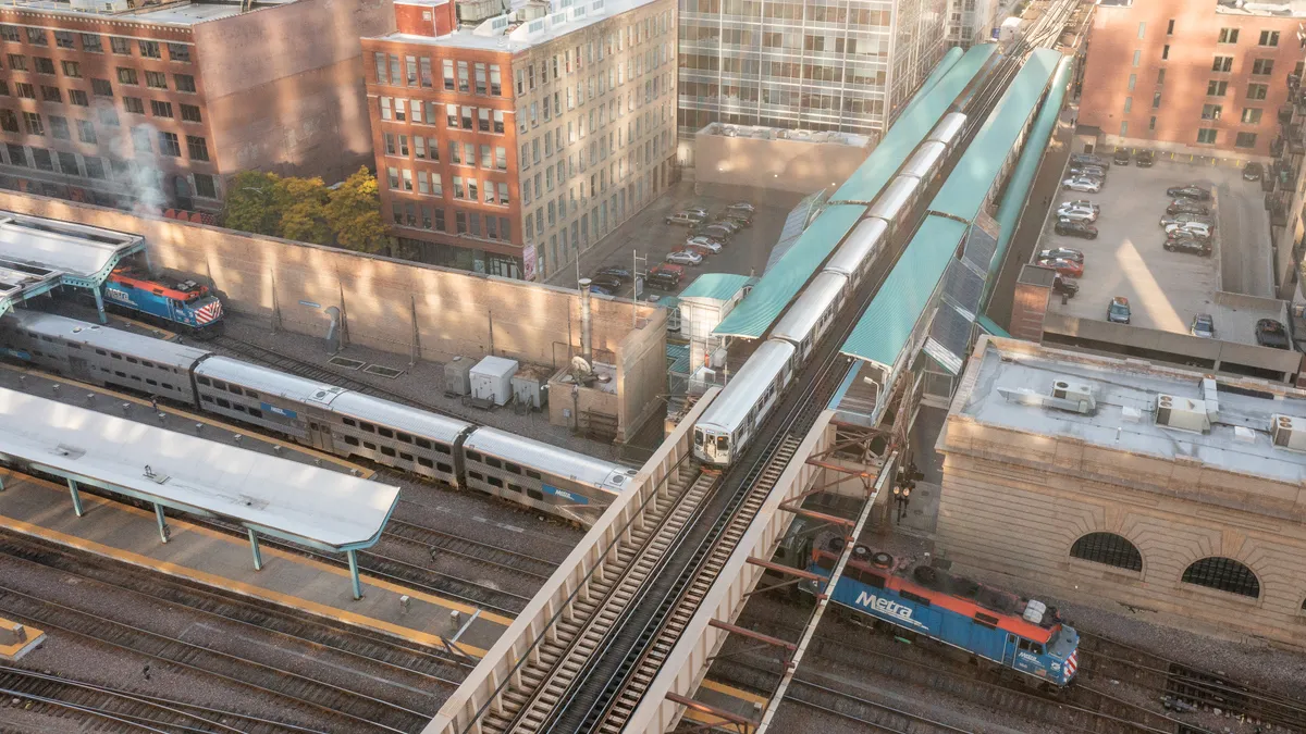 A view looking down in Chicago at an elevated train crossing from top to bottom over city streets and a railroad station with a Metra commuter train moving left to right.