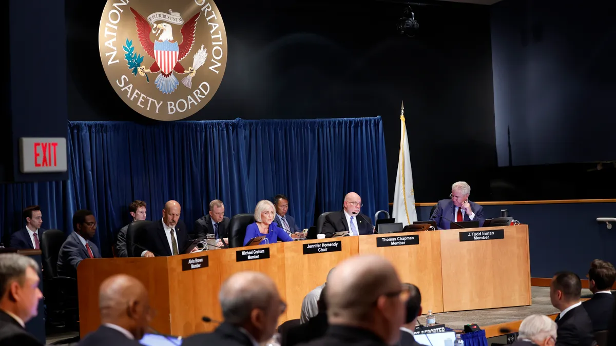 A room full of people with the National Transportation Safety Board sign hanging above them on the wall behind them.