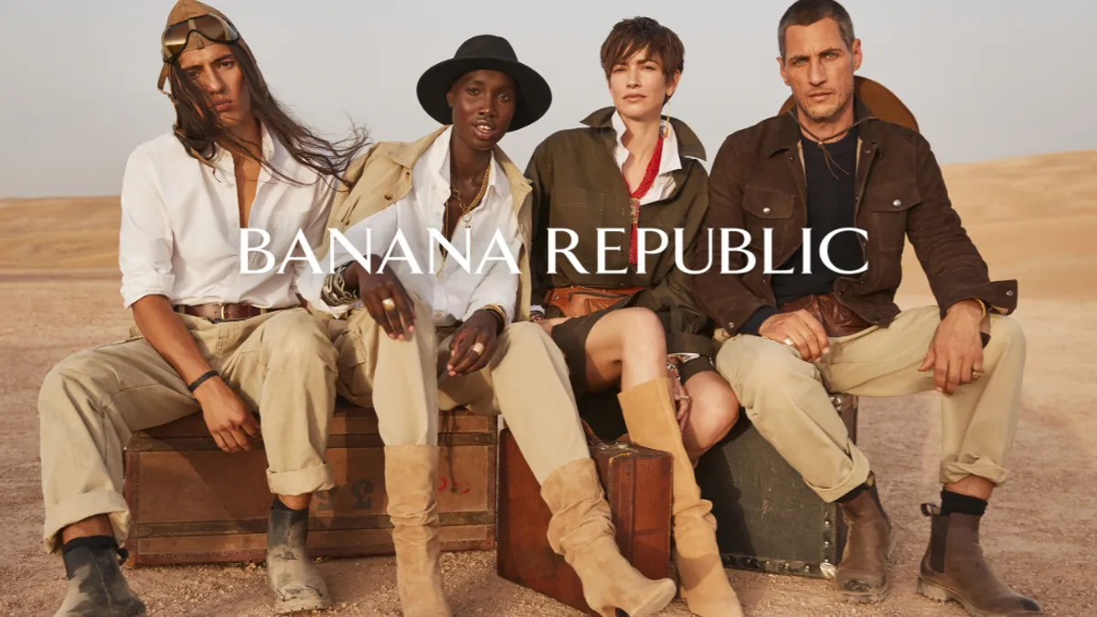 A diverse group of four people, wearing boots and khakis, sits on vintage suitcases in the desert.