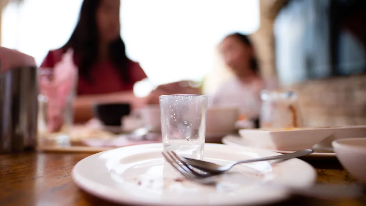 An empty plate is in focus at a table. In the blurred background, an adult and child are sitting at the same table.