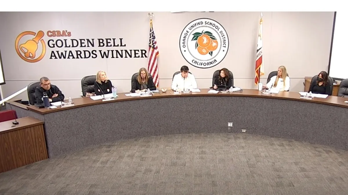 Seven adults are seated at a dias in the front of a meeting room. The school district seal is on a wall in the background.