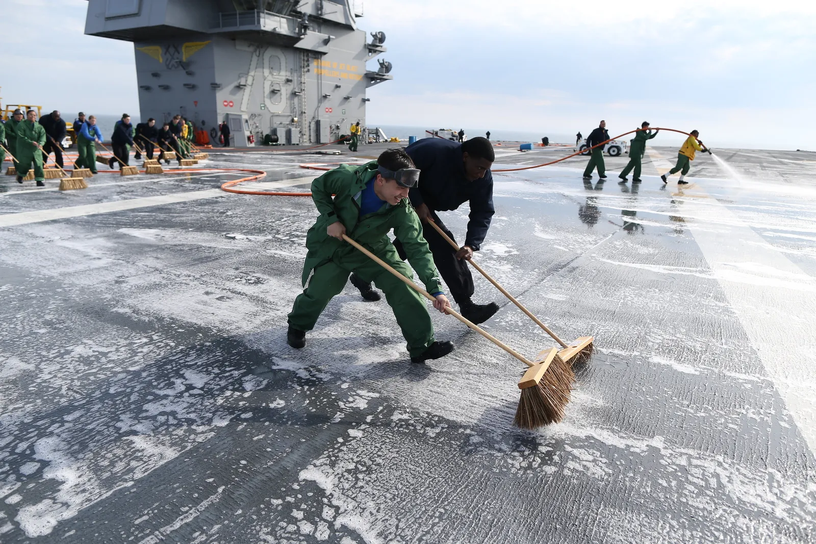 Two crew members scrub firefighting foam off the open deck of a naval vessel