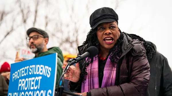 A person wearing a black hat and purple jacket and scar speaks into a podium microphone on a winter day