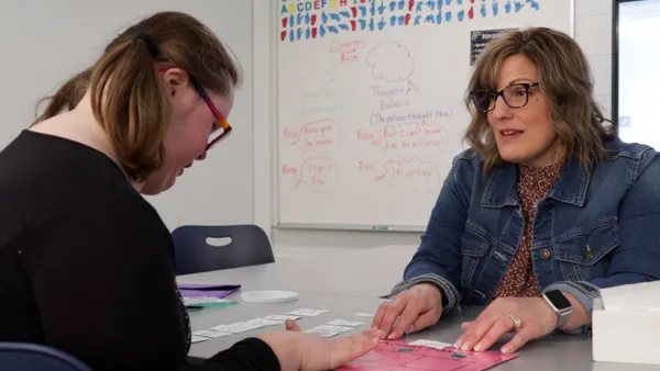 A teacher sits across a table from student as they work together in a classroom in front of a whiteboard.