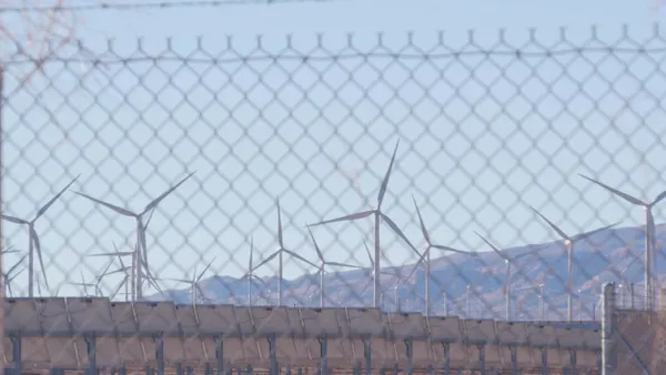 Rows of wind turbines and solar panels viewed through a chain-link fence, with mountains visible in the background under a pale blue sky.
